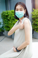 A pregnant Asian mother in a long dress is wearing a mask and thumbs up after getting vaccinated at...
