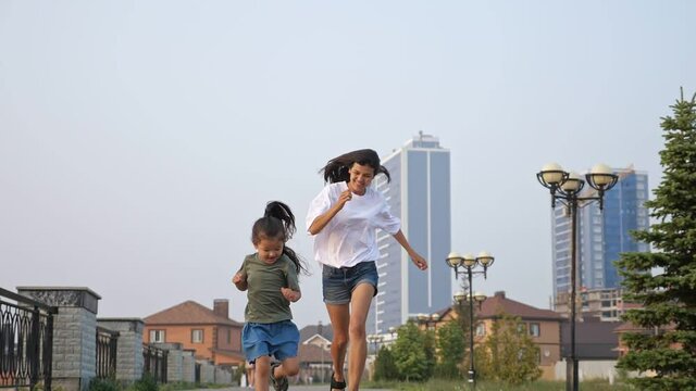 Smiling little Asian girl and long haired mother in shorts run along empty city embankment against highrise buildings at sunset time slow motion
