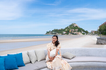 happy Asian woman tourist sit and holding pillow on sofa with blue sea beach view and mountain at Huahin, Thailand, girl relax on vacation in summer