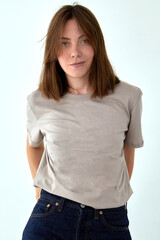 Fototapeta premium Positive female model wearing gray t shirt and jeans standing with hands behind back against white background and looking at camera