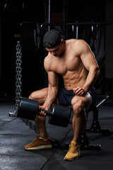 Fototapeta na wymiar an athlete with a naked torso in the gym sits on a gym trainer, holds a large black dumbbell in his hand and pumps muscles