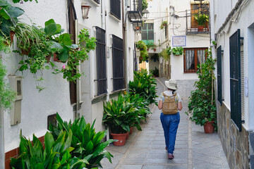 Fototapeta na wymiar Woman with a hat and a small backpack walking down a narrow street with many flowerpots, typical of the towns of Andalusia (Spain)