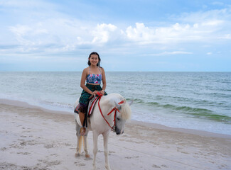 happy Asian woman tourist in beachwear riding white horse on sand beach at Huahin beach with blue sea and cloudy sky, relax on vacation in Thailand 