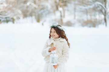 Little girl enjoys the snowfall. Little girl in a wreath holding a Christmas lantern in her hands. Christmas background.