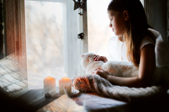 A cute kid girl with a white cat at the window with candles, a child in knitted clothes. Autumn mood and cozy art photo, long beautiful hair.