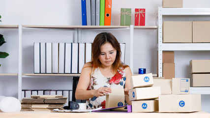 Fototapeta na wymiar Asian female small online selling business entrepreneur sitting using tape packing product package in brown cardboard box on table at home office full of empty shipment boxes and shockproof bubble