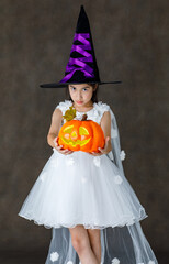 Portrait close up studio shot of Asian cute little girl in witch dress costume with tall hat stand smile look at camera hold orange pumpkin in hand ready to play trick or treat on Halloween festival