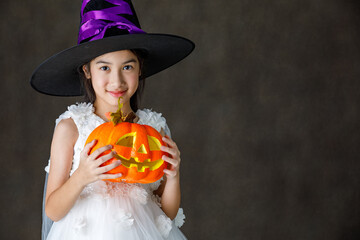 Portrait close up studio shot of Asian cute little girl in witch dress costume with tall hat stand smile look at camera hold orange pumpkin in hand ready to play trick or treat on Halloween festival