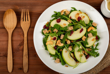Fresh green apple slice salad with rocket leaf, oat granular, dried cranberry, and cashew nut placed in white dish on the dark wooded table. The dish appeared on the right side of the picture