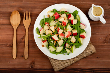 Healthy vegetarian rocket salad with dices apple, and chop almond in white dish place on sackcloth on wooded table.  Spoon fork and oil salad dressing cup beside. Two rocket leaf used as decoration
