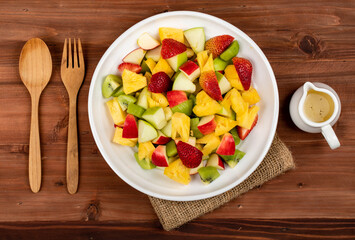 Mixed fruits salad including strawberry, kiwi, apple, and pineapple in white dish place on sackcloth on wooded table.  Utensil spoon and fork and oil salad dressing cup beside