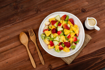 Mixed fruits salad including strawberry, kiwi, apple, and pineapple in white dish place on sackcloth on wooded table.  Utensil spoon and fork and oil salad dressing cup beside