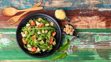 Rocket salad with tomato, crouton, herb roasted chicken in black dish together with wooden utensil...