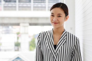 Portrait of an asian beautiful and adorable business woman wearing casual striped pattern shirt, smiling and standing in front of the window in the indoor office with background of cityscape outside.