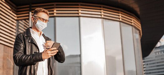 Handsome man in a protective mask on his face with glasses with a smartphone on the street of a big city. Businessman talking on the phone on urban background