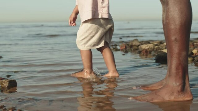 Low-section slowmo shot of unrecognizable baby boy stepping in water at beach with bare feet holding African-American father by hand