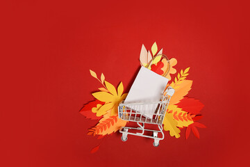 autumn shopping, leaves and and white shopping bag in grocery trolley on red background. Concept Autumn sale. copy space
