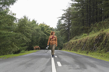 A young woman walking on the asphalt road surrounded by dense trees under a cloudy sky with brown cows in the background