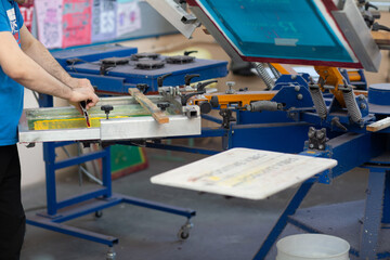 Serigraphy silk screen print process at clothes factory. Frame, squeegee and plastisol color paints.