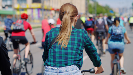 Fototapeta na wymiar Young woman wearing plaid shirt and denim shorts riding bicycle with crowd of cyclists. Contestants of sports event in city. Concept of healthy lifestyle