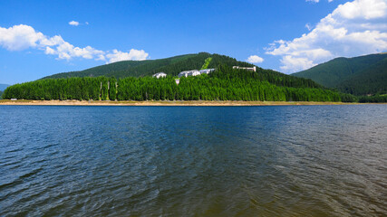 Vidra lake with its blue, clear and fresh waters in a summer day. Waves are washing the grassy shores and wild pine forests are surrounding the abandoned, old resort. Carpathia, Romania.