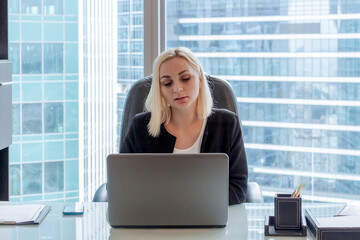 Beautiful caucasian woman with blond hair sits on chair in office and works using laptop. Blurred...