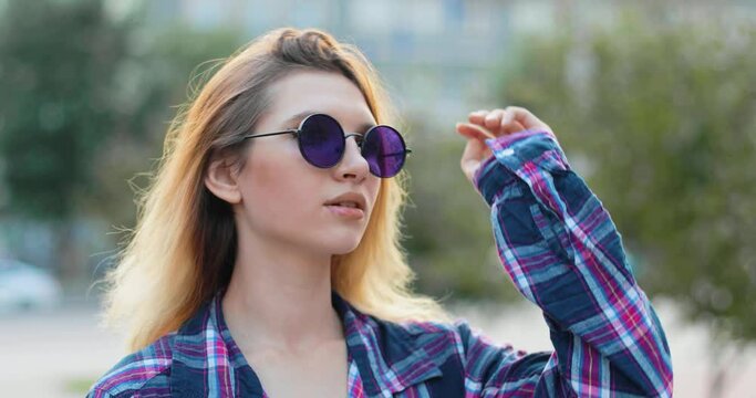Portrait of a young girl in round sunglasses. The blonde is standing in the center of the city and is looking for someone. She looks a little confused and lonely.