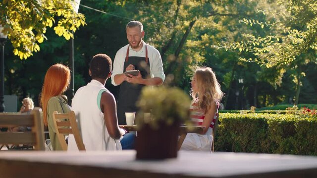 A further back view of a beautiful park with lots of tables, a sexy waiter with a beard and blonde hair is taking orders from a table with three attractive women