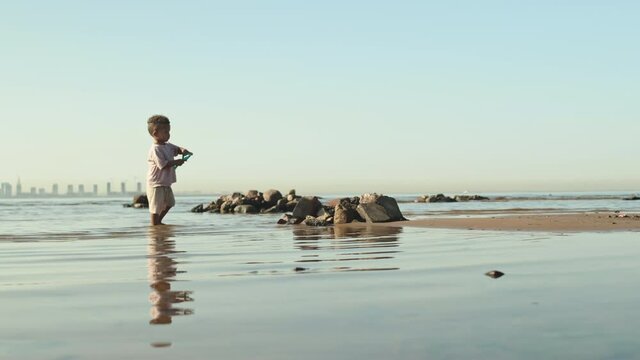 Tracking slowmo shot of cute African-American toddler boy with shovel in hands playing with sand together with his father spending summer day at seashore