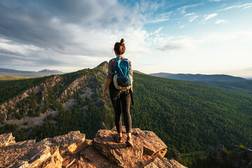 A young tourist girl with a backpack enjoys the sunset from the top of the mountain. A traveler on...