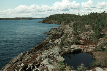 Water, forest and rocks. Wide view of scenic long craggy coastline with dark volcanic rocks of wide blue lake on sunny summer day with white low heavy clouds around rugged riverscape