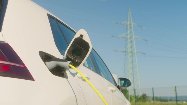 Handheld shot of a smiling Caucasian young woman unplugging electric car charger at a charging station near electric power plant, in the background visible transmission line