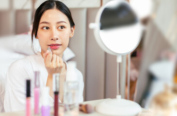 Young asian woman with towel on head applying facial scrub while looking in mirror, blurred foreground.Enjoy spa cosmetics treatment procedure, morning care hygiene concept.