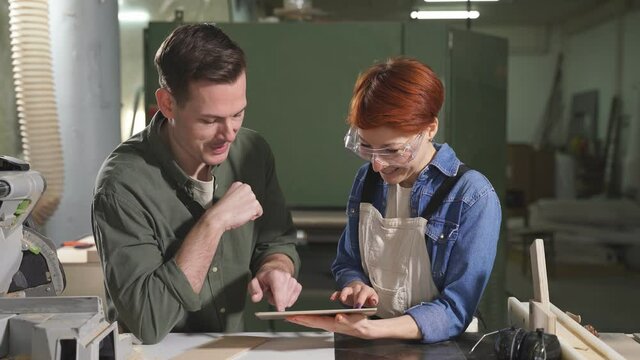Two pleasant colleagues joiners using digital tablet while working in workshop, professional carpenters discussing working process, what to do, handmade and carpentry concept