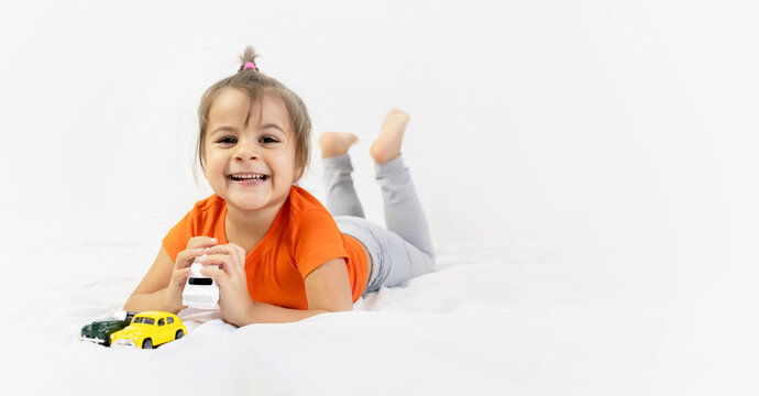 Little girl playing with white toy car. Sitting on the white bedsheet. Smiling.