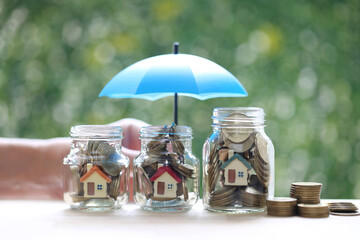Protection,Model house and gold coin money in the glass bottle with hand holding the umbrella on natural green background, Finance insurance and Safe investment concept