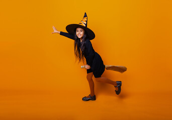 Little girl dressed as a Halloween witch in a black dress and hat flies on a broomstick on a yellow...