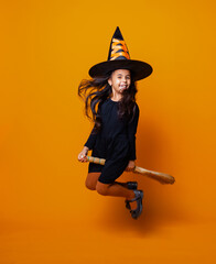 Little girl dressed as a Halloween witch in a black dress and hat flies on a broomstick on a yellow...