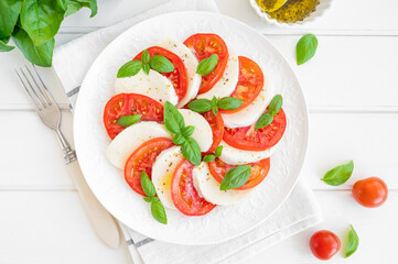 Classic caprese salad with mozzarella cheese, tomatoes and basil on a white plate on a wooden background. Top view, copy space.