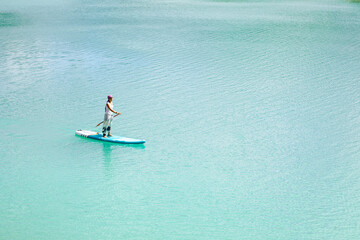 A girl in a dress floats on a glanders board on a pond with bright turquoise water. Warm summer day for travel. 