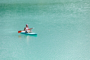 Fototapeta na wymiar A girl in a dress floats on a glanders board on a pond with bright turquoise water. Warm summer day for travel. 
