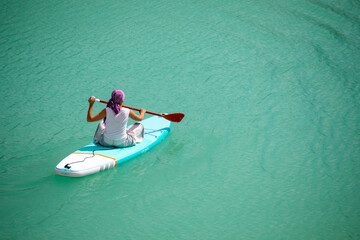 Fototapeta premium A girl in a dress floats on a glanders board on a pond with bright turquoise water. Warm summer day for travel. 