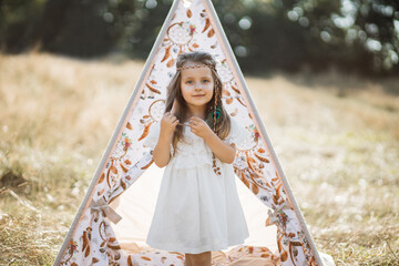 Outdoor summer shot in native american indian style. Portrait of cute little blond girl, wearing white dress and feather accessories in hair, looking at camera, standing near boho tee pee wigwam © Sofiia Shunkina