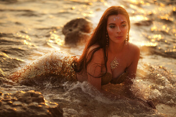 Beautiful fashionable mermaid with red hair holding shell sitting on rock near sea - obrazy, fototapety, plakaty