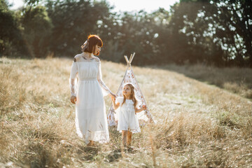Happy little girl walking outdoors in field with her mom. Woman and daughter are dressed in boho native american's style. Wigwam on the background. Happy childhood, summer holidays. Copy space