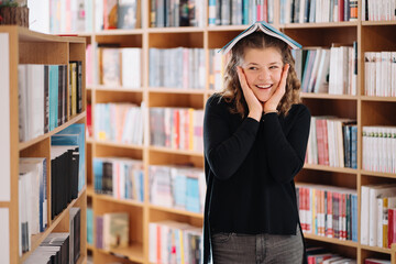 Portrait of funny smiling Caucasian teenager girl putting and holding a book on her head and looking at camera, bookshelfs background.