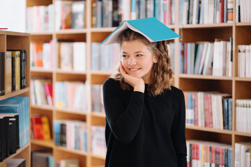 Portrait of funny smiling Caucasian teenager girl putting and holding a book on her head and looking at camera, bookshelfs background.