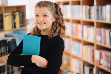 Happy tenage girl or smiling student holding a blue book wih copy space among many books in library - People, knowledge, education and school concept