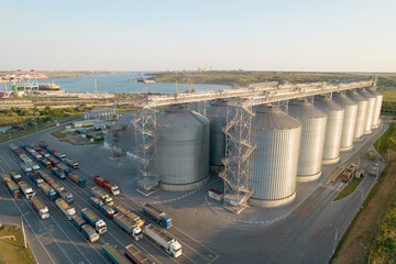 Grain terminals of modern sea commercial port. Silos for storing grain in rays of setting sun. Many trucks are waiting in line for unloading in port harbor, top view from quadcopter. Logistics, trade