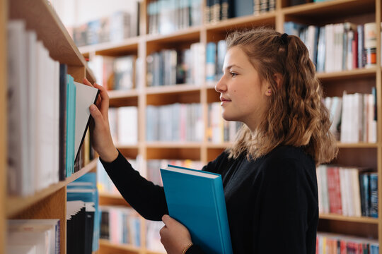 Happy tenage girl or student taking book from shelf in library - People, knowledge, education and school concept